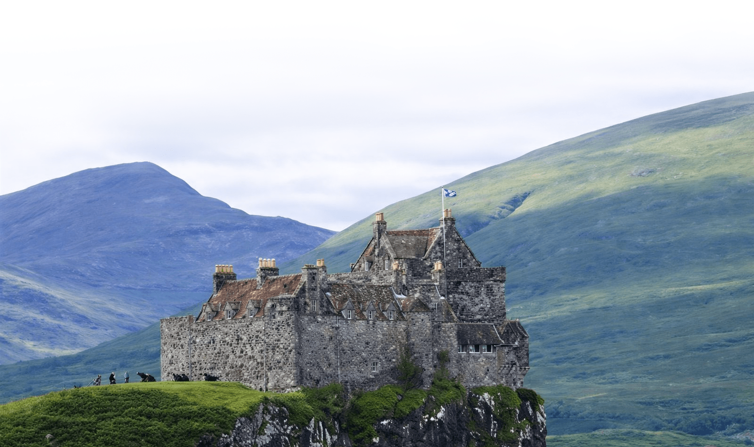 Stone castle perched on a grassy hilltop amid lush green mountains. The scene conveys a sense of history and solitude under a clear sky.